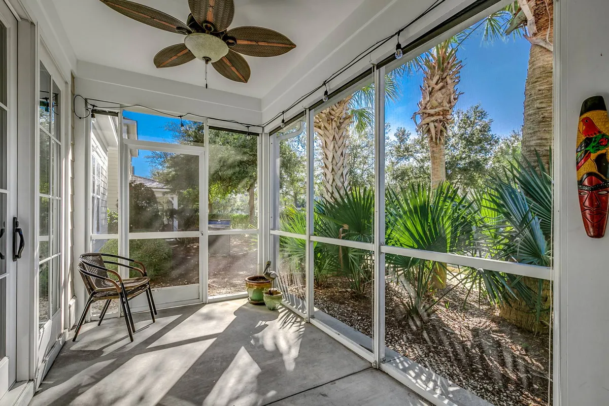 A tranquil screened porch with ceiling fan, potted plants, and decorative touches, offering views of palm trees and the outdoors. The space is perfect for socializing, enjoying nature, or savoring a quiet moment with a coastal breeze.