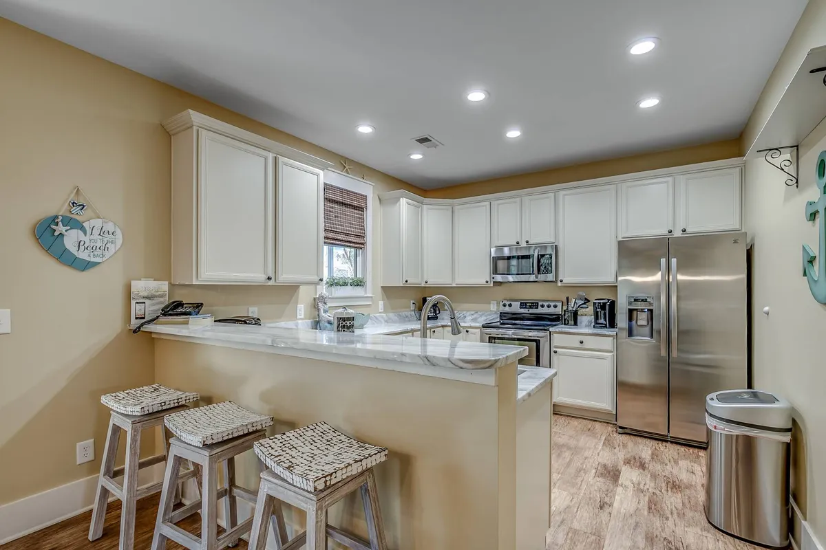 A beautifully appointed kitchen with a large island and stools, stainless steel appliances (including a dishwasher), and a window with shades allowing soft daylight to filter in. The island serves as both a prep area and a casual gathering spot, making the kitchen the hub of activity.