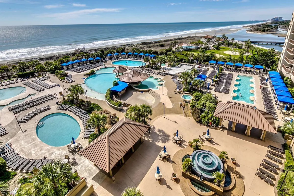 A sprawling outdoor pool complex stretches toward the beach, surrounded by swaying palm trees, landscaped gardens, and a variety of lounge chairs and poolside cabanas. The scene captures both the lively energy of people swimming and relaxing as well as the resort's beachfront location.