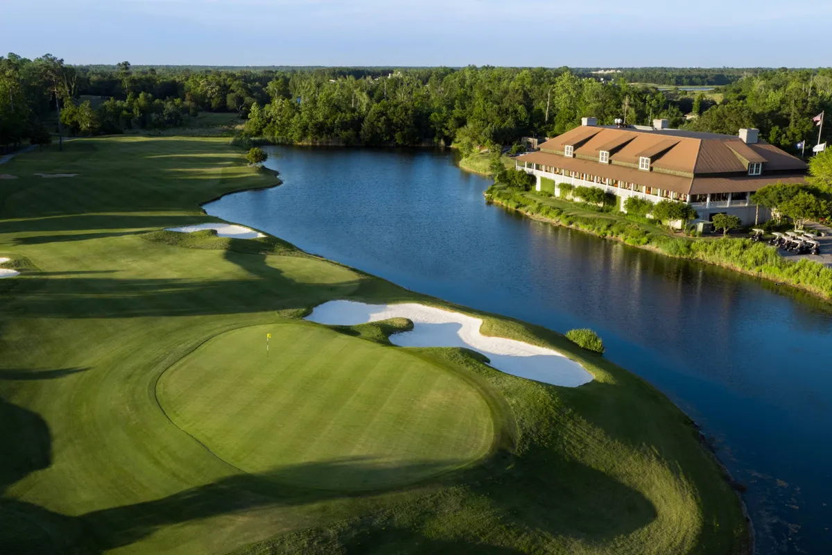 An expansive view of a beautifully manicured golf course bordered by a tranquil lake and anchored by a stately clubhouse. Sand traps add interest to the rolling green landscape, while trees and open skies convey a resort-like, leisurely ambiance.