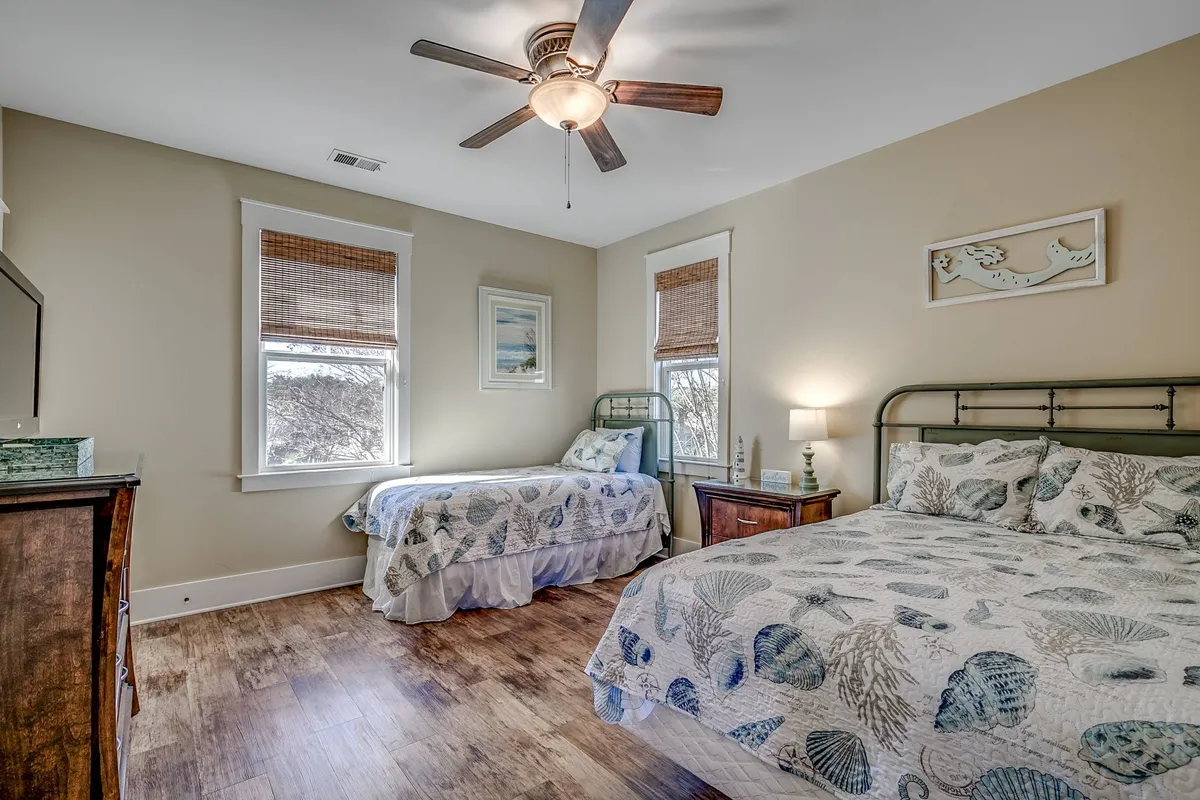 A welcoming guest bedroom featuring two beds adorned with sea-themed bedding, complemented by a ceiling fan overhead, a large window that bathes the room in natural light, and a sturdy wooden dresser. The decor evokes a relaxed, beach-inspired vibe, linking the interior to the coastal experience outside.