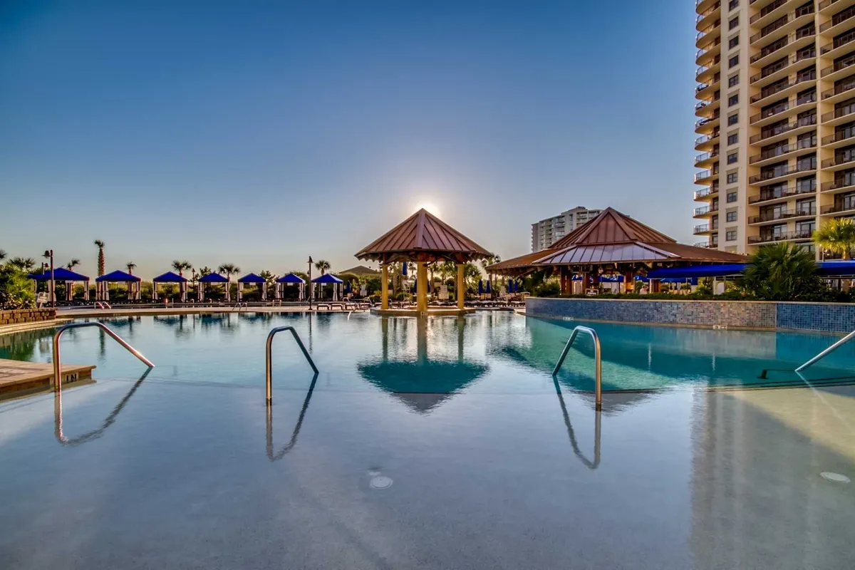 A tranquil pool scene is framed by thatched-roof cabanas and tall palm trees, all bathed in the soft glow of sunset. The image evokes a sense of calm, exclusive luxury, and tropical relaxation just steps away from the main accommodations.