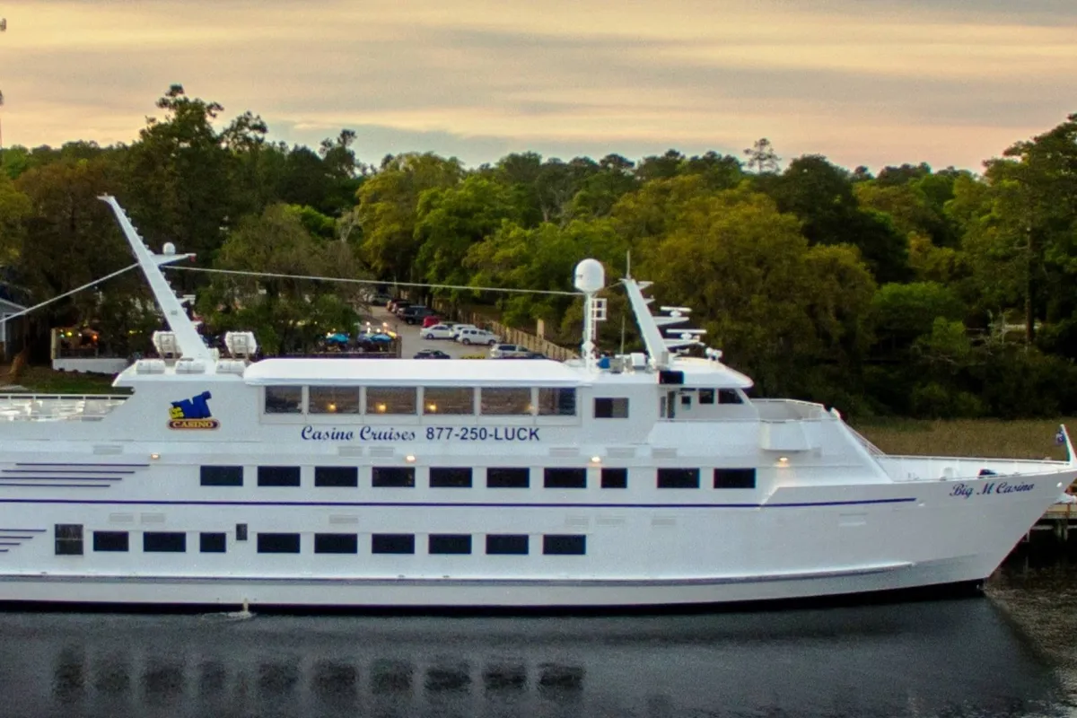 A grand white cruise ship docks near a casino featuring a distinctive logo, surrounded by lush greenery. The scene suggests excitement and a spirit of adventure, with opportunities for dining, socializing, and entertainment onboard.