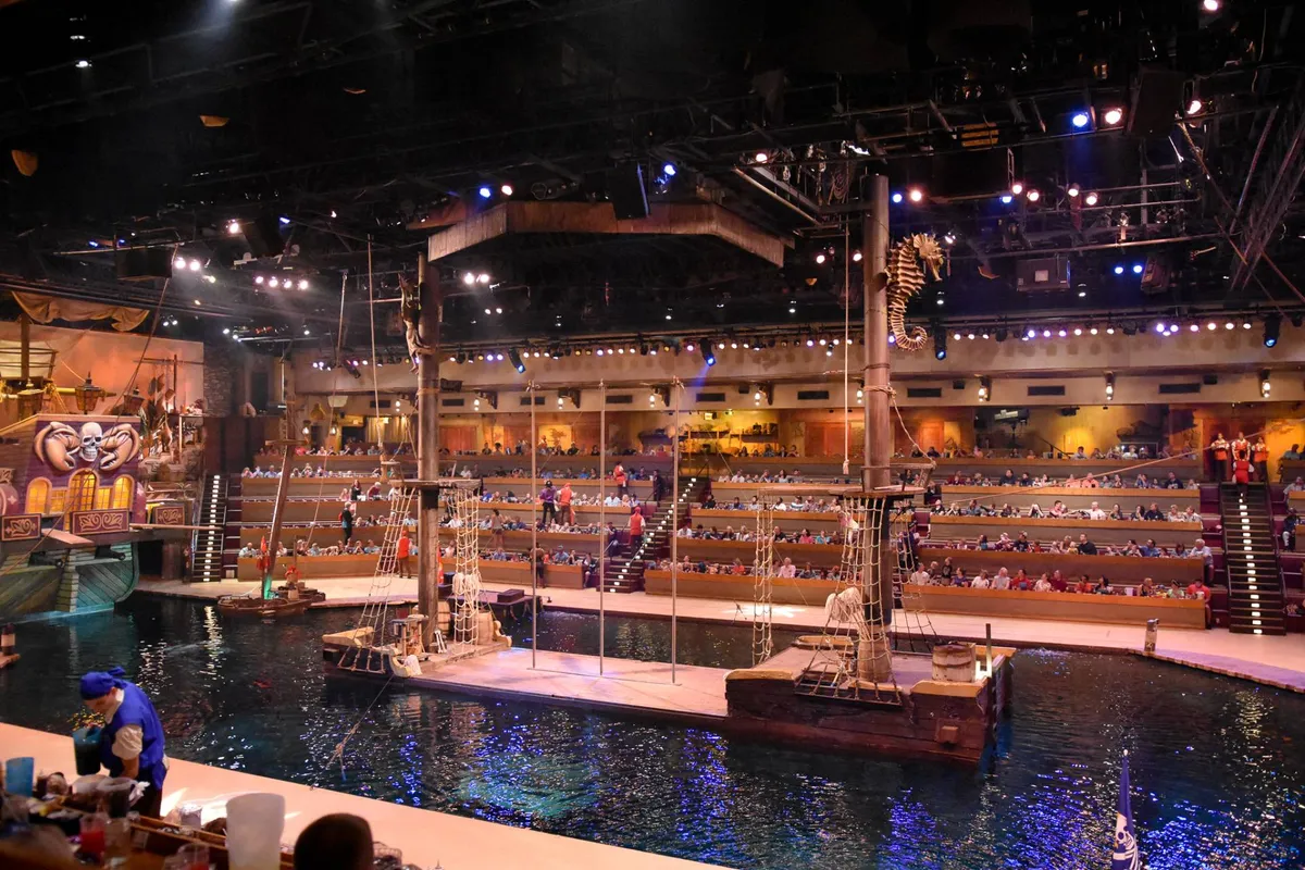 A vibrant interior view of a theatrical venue featuring an elaborate ship set, water stage, and audience seating. The energetic mood and immersive design hint at lively performances and unique dinner show experiences.